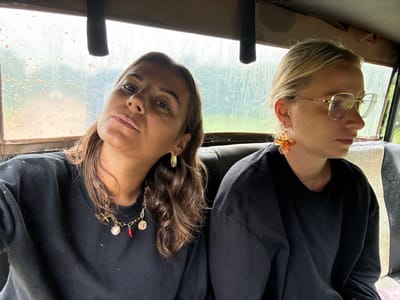 Two women pose indoors, the image presented upside down. The woman on the left has blonde hair, glasses, and large, flower-shaped earrings. She wears a dark sweater. The woman on the right has shoulder-length brunette hair, gold hoop earrings, and an embellished gold chain hanging from her sweater. Both women gaze directly at the viewer, their faces positioned toward the bottom of the frame. A grimy window with exterior views occupies the foreground.