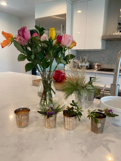 A kitchen countertop in Rye, New York (latitude: 40.952, longitude: -73.708) with white cabinets above and a white countertop, displays a bouquet of assorted roses in a clear glass vase, four small silver cups filled with sprigs of green foliage and purple flowers, and a glass vase containing a cluster of dried grass-like foliage. The background reveals a partially obscured sink with a faucet and a stainless steel range. The lighting is bright. The image appears to have been taken in a modern, well-lit kitchen.