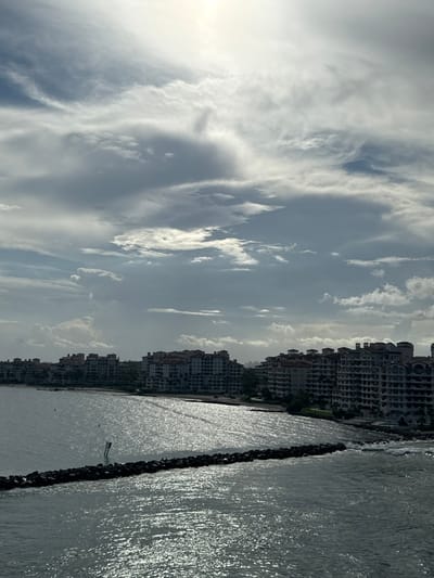 The image displays a coastal panorama identified as the waterfront of Fisher Island, Miami-Dade County, viewed from the Government Cut channel in Miami Beach, United States. The upper half of the frame is occupied by a partly cloudy sky, featuring a bright, diffused light source, presumably the sun, in the upper-right quadrant. The lower half depicts a body of water with extensive surface glare and shimmering reflections. Along the horizon, a continuous line of multi-story residential buildings, characteristic of condominiums on Fisher Island, are visible. These structures feature light-colored exteriors and dark, possibly terracotta-tiled roofs, appearing partially shadowed against the water and sky. A prominent dark, rocky breakwater extends horizontally into the water from the right mid-ground, showing wave interaction on its seaward side. A single white and black navigational marker stands vertically in the water near the breakwater. No people are discernible in the scene. The overall environment suggests a sunny, yet partly overcast day.