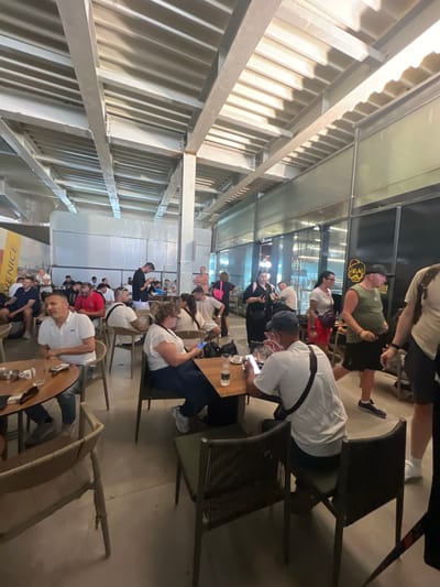 The image depicts an indoor seating area, possibly a cafe or restaurant, with a modern industrial aesthetic. The ceiling is characterized by exposed metal beams and corrugated metal panels. Fluorescent lighting is visible, casting a bright glow on the scene. A large window or glass wall lines one side of the space, offering a view of another part of the interior, which appears to be a bar or service area.
Numerous patrons are seated at tables and walking around. The individuals are dressed casually, suggesting a relaxed atmosphere. Some are engaged in conversation, while others are focused on their mobile phones. The furniture consists of tables, some round and others rectangular, with chairs that appear to be made of metal and woven material.
Objects on the tables include cups, saucers, bottles of water, and mobile phones. The floor is a neutral-toned concrete or tiled surface. The overall impression is one of a busy, contemporary public space. The location is identified as Kruja, Albania, and the establishment appears to be a bar called "Bistrot BEER".
