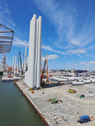 The image depicts a sunny day in the city of Esbjerg, Denmark, with a clear blue sky dotted with wispy white clouds. The scene is a port facility where components for wind turbines are being manufactured and prepared for transport. In the foreground, several large, white cylindrical structures, likely wind turbine towers, are lined up vertically on a dock. To the left, a body of water with gentle ripples laps against the reinforced concrete dock.
Further back, a variety of construction and heavy lifting equipment is visible. Several cranes, including a prominent yellow one and a tall green one, are positioned around the site. Numerous smaller, cylindrical sections, also appearing to be parts of wind turbines, are laid out horizontally on the ground in the middle and background of the image.
A barge, with "Siemens" branding visible, is docked on the left side of the image, alongside shipping containers. The dock area is a mixture of paved and gravel surfaces. Various construction materials, including stacks of green crates and concrete barriers, are scattered across the ground. In the far background, the cityscape of Esbjerg can be seen, with industrial buildings and structures under the bright sky. A few small figures of workers are present on the dock, engaged in their tasks. The overall impression is one of a busy industrial site undergoing significant construction activity.