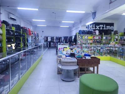 The image depicts the interior of a retail store in Ushtobe, Kazakhstan. The store appears to be a general goods or convenience store, with various items displayed on shelves and in glass display cases. In the foreground, a green, oval-shaped ottoman is visible. To the left, glass display cases line the wall, showcasing various items, possibly accessories or personal care products. Further into the store, shelves are stocked with bags, backpacks, and clothing items such as shirts and jackets. In the center aisle, tables are set up with more merchandise, including what appear to be cleaning supplies, toiletries, and household items. A banner with the text "MixTime" is prominently displayed on the back wall. The store is well-lit by fluorescent ceiling lights. No people are visible in the image. The overall environment suggests a functional retail space.