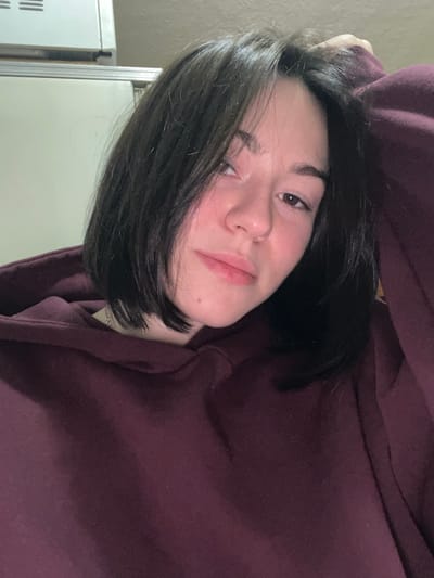 A young woman with dark brown hair styled in a short bob, wearing a maroon hooded sweatshirt, gazes towards the camera with a slight smirk. Her complexion appears fair with rosy cheeks. Part of a white appliance, possibly a refrigerator or oven, is visible in the background on the left side of the frame. The overall image is angled, creating a casual and relaxed vibe.