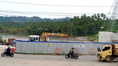 In Beunyot, Indonesia, a construction site is depicted along a river. A large yellow crane, labeled "XCMG," is positioned behind a gray, corrugated metal fence. To the left, a white truck cab is visible. A person wearing a blue jacket and helmet rides a white and black scooter on a dirt road. Further to the right, a yellow truck is parked, and another person wearing a red shirt and helmet rides a black scooter. Behind the fence, a dense forest of palm trees covers rolling hills. A river flows in the background. The sky is overcast.