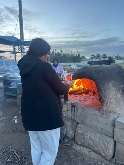 A woman in a black coat and white pants stands in front of a large outdoor oven with flames visible inside. She holds a phone, possibly taking a picture. Another person wearing a white jacket and black head covering sits nearby. The oven appears to be made of concrete blocks and stones. In the background are trees, vehicles, and an overcast sky. The ground is dirt and scattered debris.