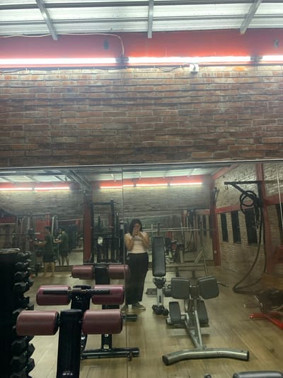 The image shows the interior of a gym in Kecamatan Kartasura, Indonesia. The gym is sparsely furnished with exercise equipment. A young woman in a white shirt and dark pants is standing near the center of the image, facing the camera, looking down at her phone. The room is dominated by a large, floor-to-ceiling mirror that reflects the gym's interior. In the reflection, a man can be seen using a weight machine in the background. The gym's ceiling is made of metal and has a single fluorescent light fixture running along its length. The walls are brick. The floor is made of wood and is covered in gym mats. There is a weight machine in the foreground with a set of red-padded seats attached to the frame. An adjustable weight bench is in the right foreground of the image, with a set of dumbbells on the floor nearby. To the right, against the back wall, is a set of weights on a stand. A rope hangs from a hook on the wall, and a set of exercise cords lie on the floor nearby. The gym appears to be empty except for the woman and the man reflected in the mirror. The woman's actions suggest she is on a break from exercising, and the man's actions suggest he is working out. The gym is clean and well-lit.