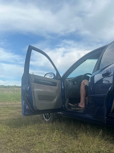 A blue car is parked in a grassy field under a cloudy sky. The driver's side door is open, revealing a person's legs and feet inside the car. The person is wearing black shorts with white stripes and black Crocs sandals. The car's interior is visible, including the steering wheel and dashboard. The background shows a line of trees and a faint structure on the horizon. The ground is covered in dry grass and dirt. The scene appears to be in a rural area outside of Ozyorny, Russia.