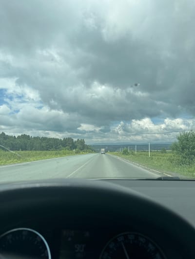 The image depicts a view from inside a vehicle, looking forward through the windshield. The road ahead is asphalt with white lane markings. On either side of the road, there are green fields and trees. In the distance, there are rolling hills and a hazy horizon under a cloudy sky. There is a white car visible on the road in the distance. The bottom portion of the image shows the dashboard of the vehicle, including a speedometer displaying a speed of 90. The scene appears to be in a rural or semi-rural area.