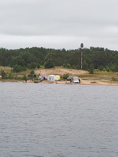 The image depicts a campsite on a sandy shore adjacent to a body of water. A line of pine trees forms the background, with a cloudy sky above. Several tents are set up on the beach, including white, grey, and a yellow and blue striped tent. Three vehicles are parked near the campsite: a dark SUV, a white camper van, and a white sedan. Several people are visible, some standing near the vehicles and tents, and at least one person is in the water. A tall, slender tower with a spherical top stands on the right side of the image, behind the trees. The overall scene suggests an outdoor recreational activity, possibly camping or fishing, in a natural environment.