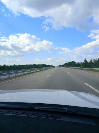 The image shows a view from the passenger seat of a car traveling on a highway. The highway has multiple lanes and is surrounded by a guardrail and a treeline. The sky is blue with scattered white clouds. The car's dashboard and hood are visible in the foreground. The scene is likely a rural or semi-rural area in Kazakhstan, given the context of Алтынды ауылдық округі.