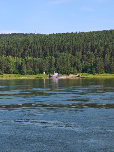 The image depicts a wide river in the foreground, with calm, rippling blue water reflecting the sky and surrounding greenery. On the far bank, a dense, green forest covers a sloping hillside. In the mid-ground, several communication towers are visible on top of the forested hill. Along the riverbank, at the base of the forest, a sandy shore is visible. A blue and white boat is partially on the shore, resting on some supports. To the right of the boat, there is a small excavator and a white car parked on the sandy bank. A sign with a white cross on a white background is also visible near the boat. The sky is clear and blue. The location is the Yenisei River near Predivinsk, Russia, a town situated on the right bank of the river.
