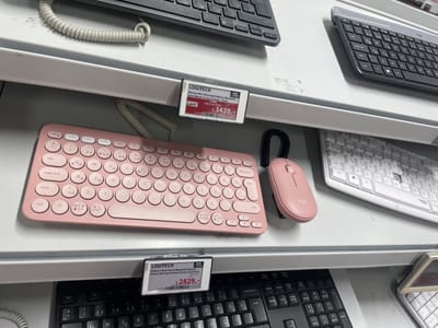 This media file shows a close-up, eye-level view of electronic devices, specifically keyboards and mice, arranged on a retail display shelf. The items are predominantly Logitech brand, with a pink Pebble 2 mouse and keyboard set prominently featured in the mid-ground. To the right of the pink set, a label indicates the price is 2,829 with the text "Pebble 2 Multi-Device Bluetooth Klavye & Mouse Seti Pudra Pembe (Türkçe Tuş Dizimi)" which translates to "Pebble 2 Multi-Device Bluetooth Keyboard & Mouse Set Powder Pink (Turkish Key Layout)". Another Logitech keyboard, the MX Keys Mini, is visible in the background with a price tag of 3,439. The setting appears to be an electronics store in Bağcılar, Turkey, suggested by the Turkish text on the price tags and product descriptions. The lighting is bright and even, suggesting an indoor, well-lit environment, possibly during daytime. The overall mood is neutral and informative, focusing on showcasing the products and their pricing.