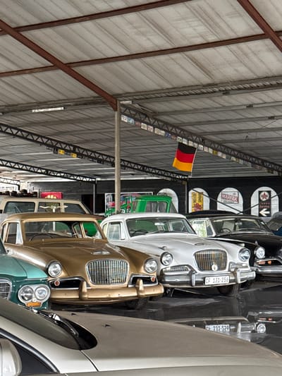 The image shows the interior of a building, likely a car museum or storage facility in Kraaifontein, South Africa. A line of vintage automobiles, including a gold one with a prominent grille, a white one with license plate "CF 131-256," and several others in varying colors (green, black, grey) are parked in close proximity. The building has a metal roof supported by metal beams and structures. A German flag is displayed, and signage with automotive branding is visible. The lighting is somewhat subdued, indicating an indoor setting.