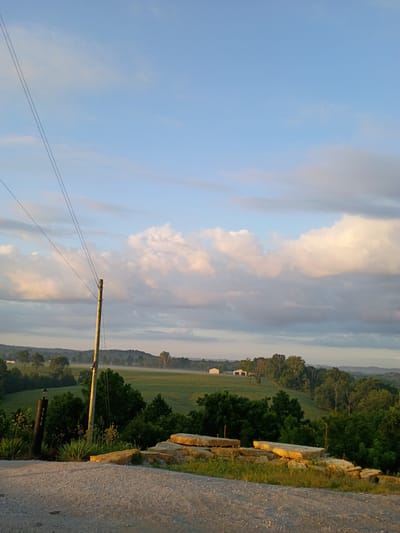 A landscape photograph depicts a rural scene in the city of Camargo, United States of America, under a partly cloudy sky at dusk. The foreground features a gravel road leading into the image, with a stack of flat, light-colored stones to the right. To the left, a dark, cylindrical post with a light fixture on top is visible, displaying the numbers "5954". A utility pole with two wires extending diagonally across the sky stands near the center.

The middle ground reveals a lush green field dotted with trees. A faint mist hovers over the field, suggesting early morning or late evening conditions. In the distance, several farm buildings, including barns or sheds, are nestled among the rolling hills. The background is characterized by a series of undulating hills covered in trees, with a hazy layer of atmosphere softening the distant view. The sky is a gradient of blue, transitioning to lighter shades near the horizon, with scattered white and pinkish clouds illuminated by the setting or rising sun. The overall impression is one of tranquil, open countryside.