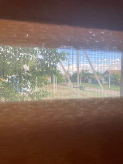 The image shows a view through a window with vertical wooden frames on the left and right sides, creating a tunnel effect. Through the window, a scene unfolds that appears to be an outdoor setting, possibly a rural area in Linyovka, Russia. The perspective is slightly skewed, and the view is somewhat obscured by the texture of the windowpane, which has small water droplets or smudges on it.
In the background, there is a clear blue sky with scattered white clouds, suggesting it is daytime, likely late morning or afternoon given the brightness. The scene beyond the window includes several structures, appearing to be small houses or buildings, amidst lush green trees. There is a path or road winding through the landscape. A greenhouse or a similar structure with white beams and translucent material is visible, stretching across the middle ground.
The foreground is dominated by the blurred textures of the wooden window frames. On the left side, the frame is a dark, rich brown, and it is speckled with small, lighter colored dots, possibly reflections or imperfections on the glass. The right side frame is a lighter, reddish-brown hue and exhibits a similar speckled texture. The foliage of trees with vibrant green leaves is visible in the lower part of the image, suggesting the season is likely summer or late spring. The overall impression is a peaceful, somewhat hazy view of a rural environment. There is no visible text in the image.