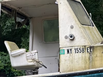 An outdoor, eye-level, medium shot captures the exterior of a weathered, off-white boat. The boat appears to be in disrepair, with visible streaks of dirt and algae on its hull. The scene is set against a backdrop of dense green foliage, suggesting an overgrown or natural environment. Prominently displayed on the side of the boat is a license plate that reads "NY 1558 E." To the left of the license plate, a smaller, teal-colored sticker with the number "19" is affixed. The boat's cabin is partially visible, revealing a white interior with a green-tinted window. A single, worn white captain's chair, also showing signs of age and use, is mounted on a swivel base and faces left. The canopy above the cabin appears torn and tattered, with fabric hanging loosely. A metal windshield wiper is attached to the upper window. The overall impression is one of neglect and abandonment.