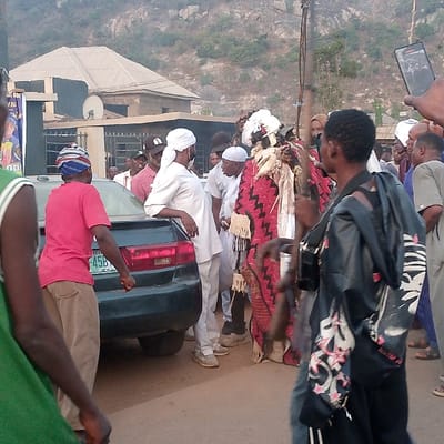A gathering of people surrounds a figure in a traditional red and white costume. Some wear white robes and turbans, indicating religious affiliation. A man in a red shirt and brown pants stands by a dark-colored car. A person wearing a black hooded jacket with white designs walks in front. Someone holds up a cell phone, possibly recording the event. A building with a satellite dish is visible in the background along with a mountain.