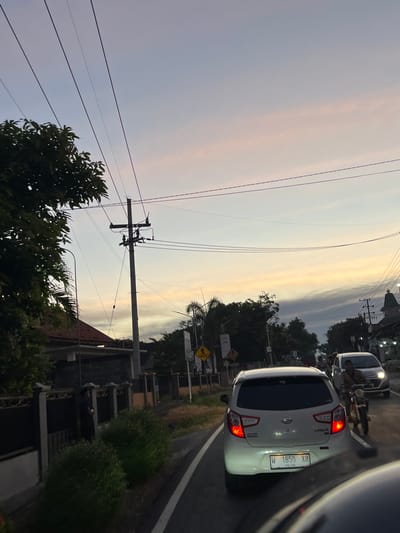 The image depicts a street scene in Purworejo, Indonesia, during what appears to be dusk or dawn due to the soft, diffused light in the sky, showing hues of pale pink and yellow. The perspective is from inside a vehicle, looking towards the right side of the road.
Prominent in the foreground and mid-ground are several cars and motorcycles, suggesting a moderate flow of traffic. A white hatchback, possibly a Daihatsu Ayla, is visible with its rear lights illuminated. The license plate displays "W 1850 XO" and "04-26", with the year "26" visible on the bottom right. Another vehicle, a silver car, is parked or moving slowly behind the white hatchback, with its headlights on. A motorcycle is also present, with its rider potentially visible.
The scene includes infrastructure such as utility poles with multiple wires crisscrossing the frame, typical of urban or semi-urban areas. Along the roadside, there are houses with visible roofs and some greenery, including trees and bushes. A yellow diamond-shaped warning sign is mounted on a pole, indicating a potential hazard.
The overall impression is a snapshot of everyday life on a street in Indonesia as the day transitions. The lighting suggests a calm, possibly cooler part of the day. No strong emotions are conveyed, but the presence of vehicles and infrastructure points to an active, populated area. The text visible is limited to the vehicle license plate.