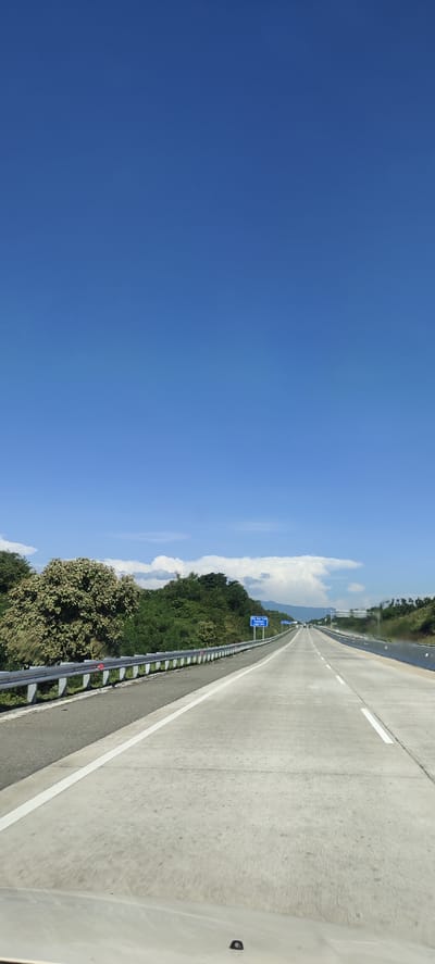 The image shows a highway with clear blue skies and a few clouds. The highway is paved with concrete and has white lines marking the lanes. There is a metal guardrail along the left side of the road, separating it from a vegetated area with trees and bushes. In the distance, there is a blue signpost on the right side of the road. The scene appears to be a daytime shot of a road in a rural or semi-rural area. There are no people or vehicles clearly visible in the foreground. The overall impression is one of travel and open road.