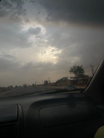 The image shows a view from inside a car, looking out the windshield. The sky is overcast with heavy clouds. On the right, a portion of the car's interior is visible, including the dashboard and vents. The road stretches out in front of the car. Buildings, trees, and utility poles line the side of the road. The image appears to have been taken in Zangon Aya, Nigeria. The road leads to somewhere. The time of day appears to be midday.
