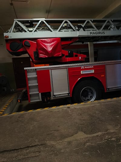 The image shows a red Magirus fire truck parked in what appears to be a fire station bay in Tukums, Latvia. The fire truck has a large, extendable ladder mounted on top. The bay has concrete flooring with yellow and black striped markings along the edge. The walls and ceiling of the bay are dimly lit and appear to be constructed of concrete or similar materials. The fire truck is the central focus of the image, with its ladder mechanism and the side compartment visible. There are no people or any other vehicles in the frame. The scene is static, with no discernible action taking place.