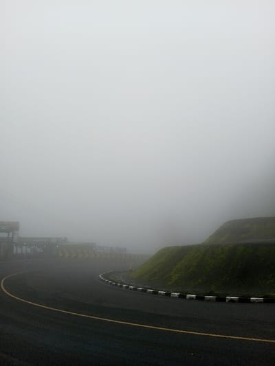 A winding road curves through thick fog, hugging a grassy hillside to the right. The road is marked with a yellow line, and a black and white curb separates it from the hill. In the distance, blurred by the fog, are structures and signs including one reading "Puncak Pass". Visibility is extremely limited, suggesting a potentially dangerous driving condition. The scene has a monochromatic, atmospheric feel.