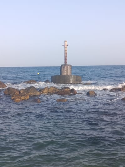 This is a photograph of a navigational marker standing in the sea off the coast of Águilas, Spain. The marker consists of a cylindrical metal structure atop a substantial concrete base, which is partially submerged. The sea is calm with gentle waves and a clear sky above. In the foreground, rocks are visible breaking the surface of the water, and a yellow buoy floats further out to sea. The scene appears to be a tranquil coastal environment.