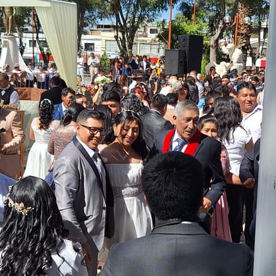 The image depicts a wedding ceremony outdoors. Many people are in attendance. The couples getting married are visible among the crowd. There are trees, buildings, and a white canopy. Large black speakers are near the crowd. Based on the location tag provided, the event appears to be in Cerro Colorado, Peru.