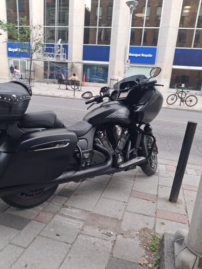 A black Indian motorcycle is parked on a paved sidewalk in front of a building with large windows. The motorcycle is equipped with saddlebags and a windshield. In the background, people are visible sitting on a bench and walking on the sidewalk. A bicycle is parked across the street. The building appears to be a bank with signage in both English and French, "Royal Bank" and "Banque Royale", suggesting the location is in Ottawa, Canada. The scene is typical of an urban environment with street furniture and pedestrian activity.