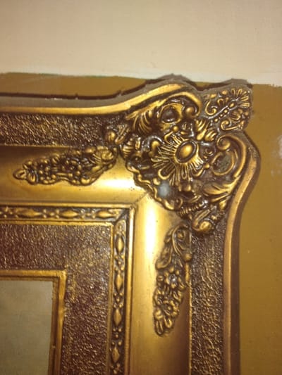 The image displays a close-up, angled view of the upper right corner of an ornate, rectangular, gold-colored picture frame. The frame features intricate relief ornamentation, most prominently a heavily sculpted floral and foliate motif at the corner, centered by an oval element. This corner motif extends along the frame's edges with further decorative scrolls and clusters of textured elements resembling berries. An inner border of the frame exhibits a granular or stippled texture, while an adjacent border presents a repeating pattern of elongated ovals or diamonds framed by parallel lines. The overall finish is a metallic gold, showing signs of dust accumulation, particularly on the topmost edge, and minor surface wear or imperfections within the decorative crevices. A blurred, light-toned, possibly reflective surface is partially visible at the bottom left, indicating the framed item. The background consists of a light cream or off-white wall section above the frame, meeting a yellowish-brown wall surface to the right. No individuals, discernible actions, or specific contextual elements are present in the image to identify the location as Ciudad de San José, Costa Rica.