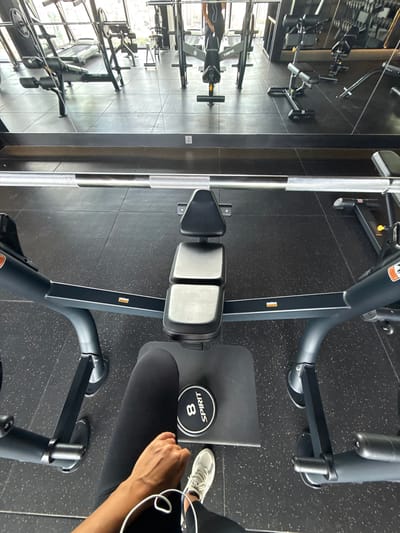 The image shows a person sitting on a weight machine in a gym. The machine is black and has a padded seat and backrest. A person's hand is visible, wearing a white athletic shoe and black leggings. A weight plate with the brand "SPIRIT" and the number "8" is on the floor next to the person. The gym has a black rubber floor with speckled gray and white flecks. In the background, there are other gym machines and a mirrored wall that reflects more gym equipment. The overall scene suggests a typical gym environment. The lighting appears to be bright, indicating it might be daytime. There is no discernible weather information. The location context is Khan Mean Chey, Cambodia.