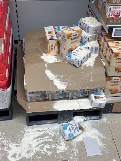 The image shows a spill of flour and related products, possibly in a retail setting, likely a grocery store, in Jestetten, Germany. Several packages of "Weizenmehl Type 405" (wheat flour type 405) and "Paniermehl" (bread crumbs) are visible, some with damaged packaging and flour scattered around them. One bag of "Weizenmehl" is torn and lying on the floor with a significant amount of flour spilled around it. The spill is on a tiled floor next to a black plastic pallet. The products appear to be on shelves or displays in a retail environment. Other products like "Spezialmehl Fur Pizza" are visible on a shelf above.