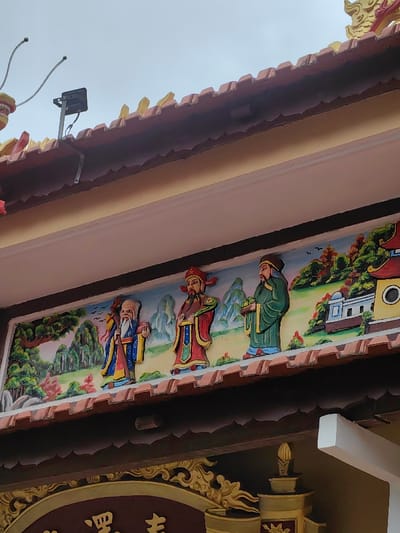 This image captures a detail of a building facade, likely a temple or pagoda, in Phú Xuân Ward, Vietnam. The scene is dominated by a vibrant, rectangular relief panel featuring three anthropomorphic figures, commonly identified as the Fú Lù Shòu (Three Star Gods).

From left to right:
The first figure is an elderly male with a long white beard and a prominent forehead, dressed in a blue robe with yellow trim. He holds a staff in his left hand and an indeterminate object in his right.
The central figure is a male with a dark beard, wearing a red robe with intricate yellow and blue patterns and a red cap. He holds a green ruyi scepter in his right hand.
The third figure is a male with a dark beard, clad in a green robe with yellow trim and a dark cap. He holds a cluster of peaches in both hands.
The background of the relief depicts a stylized landscape with green mountains, trees, and small, traditional buildings under a light blue sky with several bird silhouettes.

Above the relief, a row of reddish-brown, undulating roof tiles is visible, surmounted by a lighter-colored eave. A black rectangular floodlight or surveillance camera is mounted on a light-colored metal bracket on the roof, with visible wiring extending from it. To the immediate left of the light, a yellow and red decorative element, possibly part of a dragon or mythical creature, with two spring-like wires protruding, is partially visible.

Below the relief, an ornate dark brown frieze features golden decorative patterns and partially visible Vietnamese script, forming part of a larger inscription or sign. Beneath this, a plain light-colored (possibly pinkish-beige) wall surface is present. The sky above the building appears overcast and light grey.