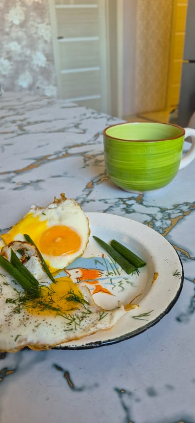 A plate of fried eggs with scallions and dill sits on a marble table. A green mug is visible to the right of the plate. The image appears to be taken indoors, likely in a home kitchen. There is no visible indication of the city of Yemelyanovo, Russia.