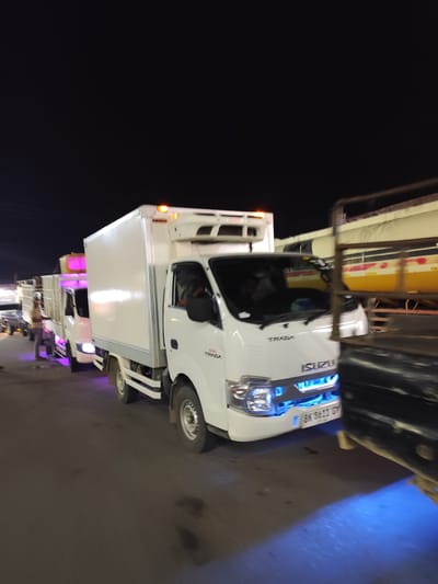 The image depicts a nighttime scene in Meunasahceubrek, Indonesia, featuring a line of white box trucks on a paved road. The sky is dark, indicating late hours.

In the foreground, a prominent white Isuzu Traga truck is visible. Its front grille clearly displays the "ISUZU" logo, and "TRAGA" is written on the cab. This truck is equipped with distinctive bright blue underglow lighting that illuminates the road surface beneath its front bumper. Orange marker lights are also active on the top front of its refrigerated box body. A license plate, partially visible as "BK 8611 GM," is affixed to its front.

Behind the lead truck, several other white box trucks are queued, some also featuring underglow lighting, such as a purple-pink hue emanating from a truck further down the line. A man is seen standing near one of these trucks, suggesting activity related to waiting or handling cargo. On the far right, a dark, indistinct object partially obscures the view. In the background, a large, yellow structure with red markings, possibly a storage tank or part of a larger vehicle, is dimly visible. The scene portrays a functional night environment, likely a loading zone or a waiting area for commercial vehicles.