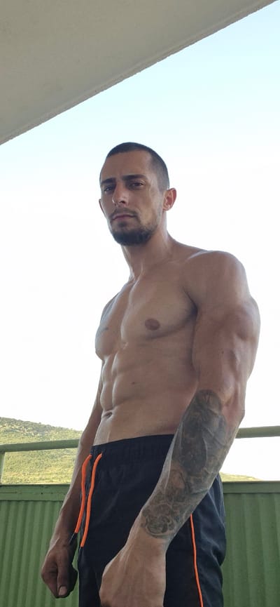 The media file shows a shirtless man with a muscular physique and tattoos on his arm. He is wearing black athletic shorts with orange trim. The image is taken from a low angle, looking up at the man against a bright sky. The man has short dark hair and a beard, and he is looking towards the camera with a neutral expression. In the background, there is a green corrugated metal wall and a hint of lush vegetation, suggesting an outdoor setting. The lighting indicates it is daytime, possibly a bright, sunny day. The orientation of the image is rotated, with the man appearing sideways. The scene appears to be set in Kaspichan, Bulgaria, based on the provided location context. There is no discernible text visible in the media file.