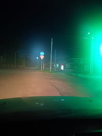 The image depicts a nighttime scene on a paved road in the city of Ponomarevka, Russia. A road sign indicating a mandatory turn left or straight is visible in the middle ground, accompanied by a yield sign. To the right of the road, a building with a sign that reads "МАГНИТ" (Magnit) is illuminated. The facade of the building has multiple windows. The road is lit by streetlights, and a bright green light emanates from the right side of the frame, likely from a light source not fully captured. The foreground shows a dark surface, possibly the interior of a vehicle. The sky is dark, with no visible celestial bodies. The overall environment appears to be a residential or commercial street at night.