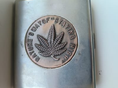 The image shows a close-up of a metal flask with an embossed design. The design is circular and features a cannabis leaf in the center, surrounded by text that reads "NATURE'S WAY OF 'STAYING HIGH'". The flask is silver-toned and has a brushed metal finish. The lighting suggests an indoor setting, possibly a desk or table. There is no indication of the location being in the city of Kalynivka, Ukraine. The image is static and there are no people or actions depicted.