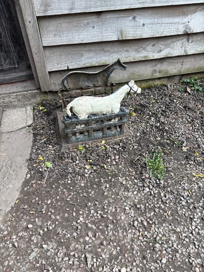 The image shows a cast iron boot scraper with a white horse figure attached, lying next to a weathered wooden wall. The surrounding ground is covered with small stones and gravel, suggesting an outdoor environment, possibly a garden or yard. The location is assumed to be Pulverbatch, United Kingdom. No people or actions are depicted.
