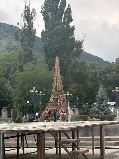 This image captures a view of a replica Eiffel Tower, a prominent landmark in Goris, Armenia. The tower, constructed from wood, stands tall amidst lush greenery, with a backdrop of rolling hills and a cloudy sky. In the foreground, a metallic structure, possibly part of a stage or exhibition, partially obscures the view. Several people are scattered throughout the scene, some seated on benches, others walking, and a few are near the base of the tower. A fountain sprays water to the left of the tower, adding a dynamic element to the composition. The overall atmosphere is serene and relaxed, suggesting a public park or recreational area.