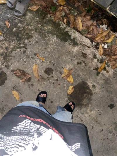 The image shows a first-person perspective of someone standing on a wet, concrete sidewalk in Banjarmasin, Indonesia. Fallen, dry leaves are scattered across the ground, along with some damp patches indicating recent rainfall. The person is wearing blue jeans and black slide sandals. Their black t-shirt, with white graphic text, is partially visible in the foreground. In the background, more dry leaves and debris are visible near a concrete edge, suggesting the location is an outdoor area, possibly near a gutter or drainage channel. The overall lighting suggests it is either evening or nighttime.