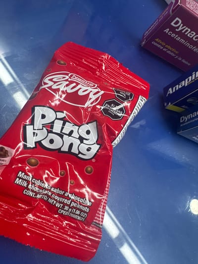The image shows a red wrapper of a "Ping Pong" candy from Savoy Nestle on a blue surface. The package has Spanish text indicating it contains milk chocolate covered peanuts. To the left of the candy are two boxes of medication: "Dynac" and "Anapi," medications commonly used to reduce fever and pain. The details on the wrapper indicate the product is manufactured in Parroquia Ocumare, Venezuela. There is no information regarding people, subjects, or actions.