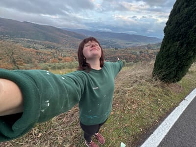 An upside-down selfie shows a woman in a green sweatshirt with white paint splatters, standing on grassy terrain. Black leggings and pink shoes are visible. Beside her is a road with a white line. The background features a tree, a landscape with hills, and a cloudy sky. Autumnal colors are prominent in the vegetation. The perspective is unusual, turning the typical landscape view on its head.