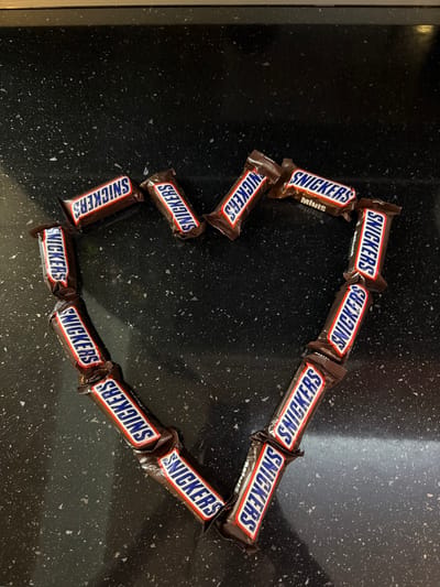 A collection of approximately sixteen Snickers mini chocolate bars are precisely arranged to form the outline of a heart shape on a dark, speckled surface. Each chocolate bar is individually wrapped in its standard brown packaging, featuring the "SNICKERS" logo in white, red-outlined letters on a blue rectangle. The text "MINIS" is visible on several wrappers, oriented in various directions as dictated by their placement within the heart's curvature. The bars are laid flat, with some slightly overlapping at their ends to maintain the heart's form. The surface beneath the candy is a polished, dark countertop, likely granite or quartz, exhibiting a fine-grained pattern of white and grey flecks against a black background. The lighting is even, illuminating the scene without harsh shadows. No individuals are present. The specific city of Yoakim Gruevo, Bulgaria, cannot be confirmed from the visual information presented.