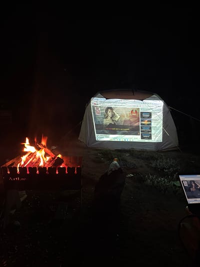 In the dimly lit outdoor environment of Sophiyevka, Kazakhstan, a scene unfolds featuring a campfire and a projected image on a tent. The campfire, positioned in the foreground, is actively burning with visible flames and embers within a metal brazier that has "ArtLine" inscribed on it. To the right of the campfire, a tent is set up, and its surface is acting as a screen for a projected image. The projected image appears to be a webpage, possibly from YouTube, displaying a woman's portrait and other video thumbnails with Russian text. To the far right, a laptop screen is also visible, displaying a similar webpage with a woman's image. The surrounding environment is dark, with some low-lying vegetation visible around the tent. The overall impression is a relaxed, evening outdoor gathering or movie-watching experience.