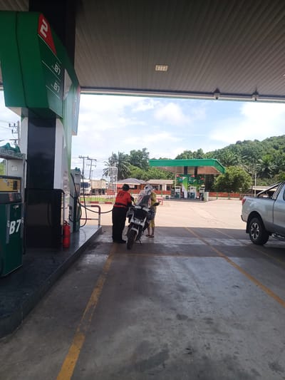 The image shows a scene at a gas station in Plai Phraya, Thailand. A gas station attendant wearing an orange shirt is interacting with a person on a motorcycle. A silver pickup truck is parked nearby. The gas station has green and white branding. In the background, lush green vegetation and a building are visible. The sky is partly cloudy.