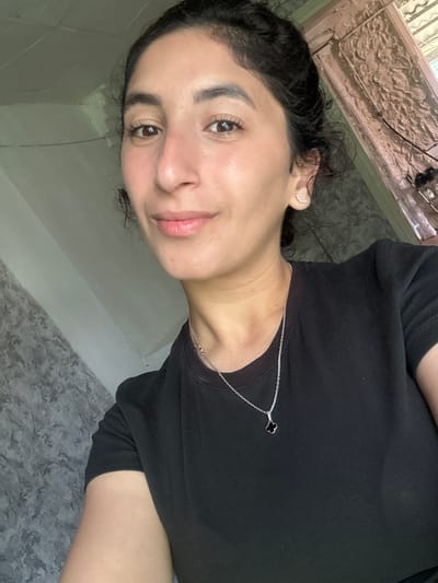 A young woman with dark hair pulled back and brown eyes is looking at the camera. She is wearing a black t-shirt and a silver necklace with a clover pendant. She has small, delicate earrings in her right ear. The background appears to be indoors, with a textured gray wall on the left and a lighter, possibly stuccoed wall on the right. Some architectural details are visible in the upper right corner. There is no indication of the city of Dondukovskaya, Russia in the image.
