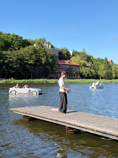 A young man stands on a wooden pier, fishing with a rod in a body of water. He is wearing a white t-shirt and black pants. The pier extends into the water, which is calm and reflects the clear blue sky. In the background, a swan-shaped pedal boat with two people is visible, as well as a grey car-shaped pedal boat with three people. Behind the water, a lush green hillside is covered with trees and buildings, including houses with red tile roofs. The scene appears to be set in a park or recreational area. The location is the lake near the city of Svetlogorsk, Russia.