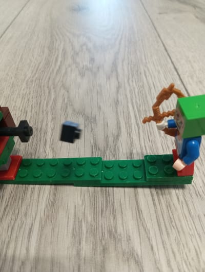This is a close-up, eye-level shot of a Lego diorama set on a wooden floor. The diorama depicts a scene with two Lego figures engaged in a simulated battle. On the left, a Lego structure, possibly a turret or weapon, is visible, featuring red, green, and grey Lego bricks. A black Lego projectile is captured mid-air, just in front of the structure, suggesting it has just been fired. On the right, a Lego figure, dressed in blue and green with a red base, is aiming a brown Lego bow and arrow. This figure appears to be the target of the projectile or is in the process of retaliating. The scene is set against a backdrop of a light-colored wooden floor with distinct grain patterns, suggesting an indoor environment. The lighting is bright and diffused, with no obvious shadows indicating a specific time of day or weather conditions. The overall mood is playful and action-oriented, typical of a Lego play scene. No text is visible in the image. The location context of Oleksandriia, Ukraine, is provided but not directly inferable from the visual elements of the image itself.