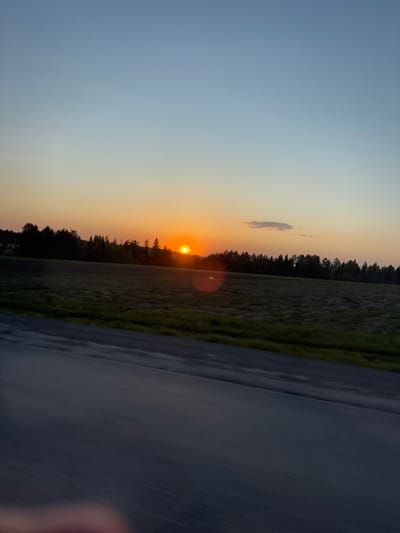 The image depicts a sunset over a field, likely taken from a moving vehicle. A road occupies the right side of the frame, its surface appearing wet. Beyond the field, a line of trees separates the sky from the ground. The sun is positioned near the horizon, casting a warm glow and creating a lens flare. A faint cloud is visible in the upper portion of the sky. The color gradient in the sky transitions from blue to orange.
There is no information to associate the image with Kamennoye, Russia.